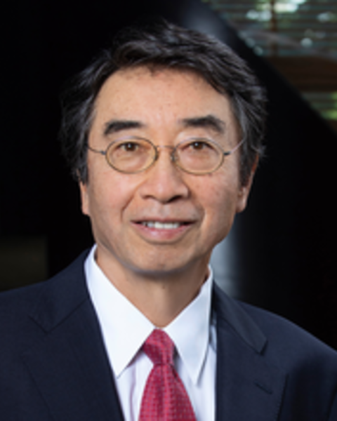 A Japanese man with glasses wearing a suit and tie smiles at the camera